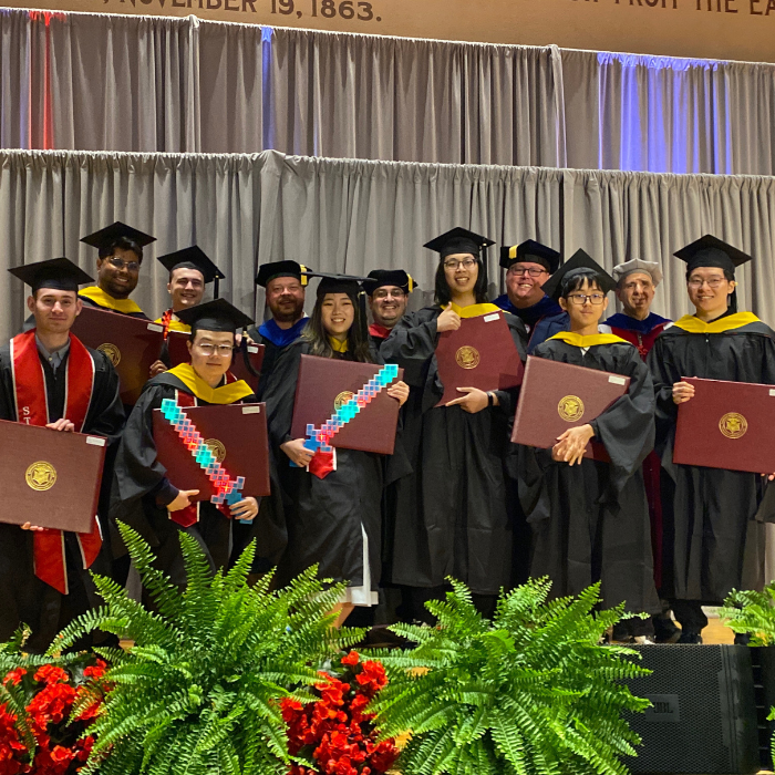 A group of men and woman pose together smiling for the camera wearing graduation regalia